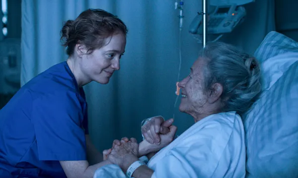 A still from the film Late Shift. A nurse comforts a grey-haired older woman who is propped up in a hospital bed.