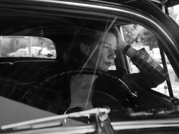 A still from the black and white film Fatherland. A white woman sitting in a car rests her left elbow on the bottom of the open window and rests her face on her left hand.