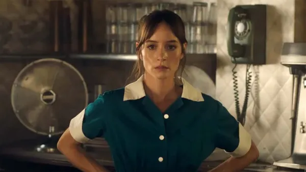 A still from the film The Last Stop In Yuma County. A white woman in a dark green waitress' uniform stands in a kitchen area with her hands on her hips, looking worried.