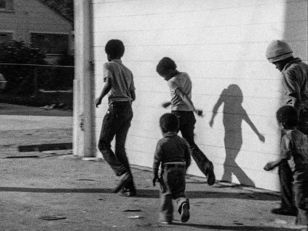 A black and white still from the film Killer Of Sheep. Five Black boys proceed cautiously around the edge of a white wall.