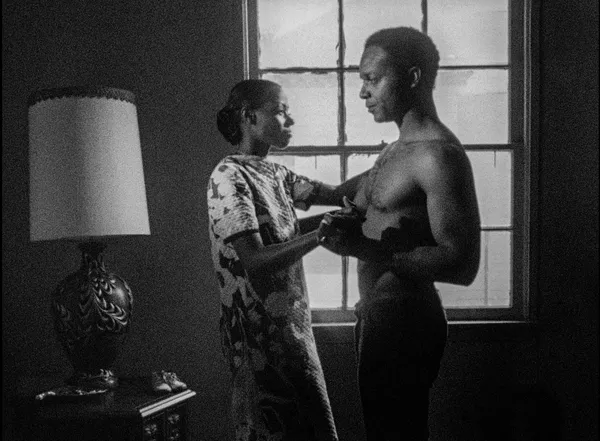 A black and white still from the film Killer Of Sheep. A Black woman embraces a shirtless Black man in front of a window.