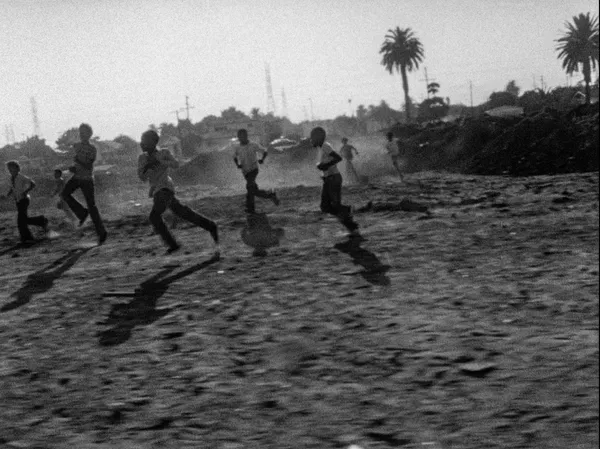 A black and white still from the film Killer Of Sheep. A group of Black boys play on a barren patch of ground.