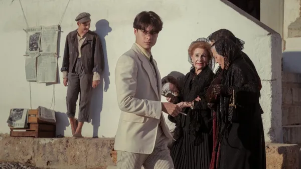 A still from the film La Bola Negra. A young white man in a pale suit flicks a cigarette as he walks through a Mediterranean town, past a thin white man in a flat cap and two white women in long black dresses.