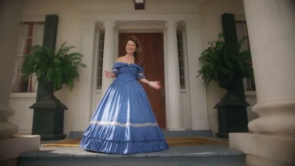 A still from the film Natchez. A middle aged white woman in an azure gown with hoop skirt stands in front of the pillared doorway of a plantation era house, raising her palms and smiling in delight.