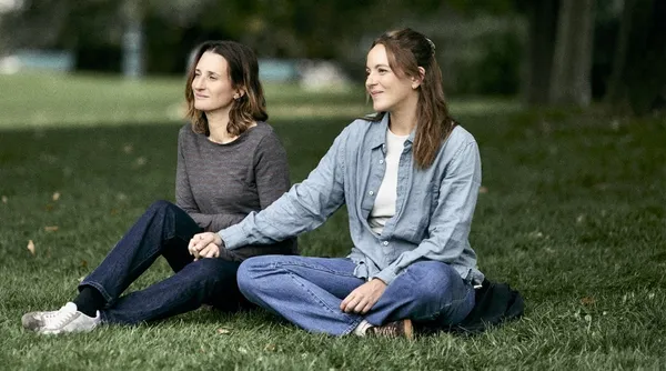 A still from the film Out Of Love. Two casually dressed white women site on the grass in a park, holding hands.