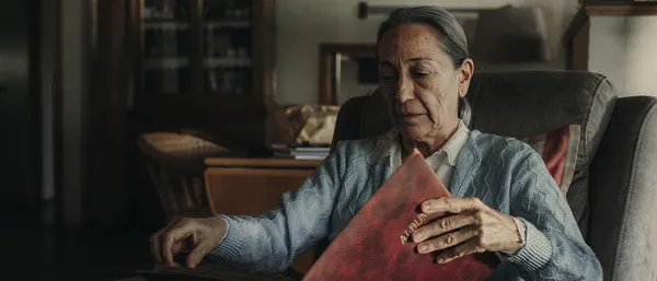 A still from the film Paris 70. An elderly white woman sits in a chair, holding a closed photograph album.