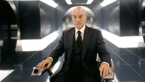 A still from the film X-men. A bald middle-aged white man sits in a wheelchair in a futuristic corridor. He has a serene expression.