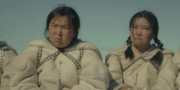 A still from the film Wrong Husband. Two Inuit women, one young and one middle aged, stand side by side under a pale sky, wearing traditional dress.