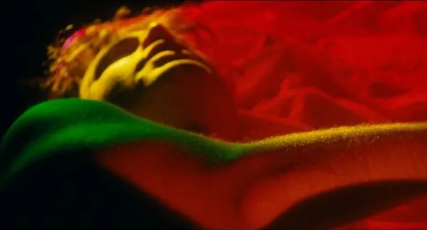 A still from the film Anything That Moves. As he lies on his back in a bed, the face and right arm of a young white man can be seen bathed in red and green light.
