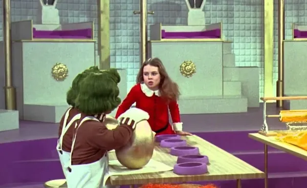 A still from the film Willy Wonka And The Chocolate Factory. An orange-skinned, green haired oompa-loompa tends to a golden egg while a white girl in a red dress dances.